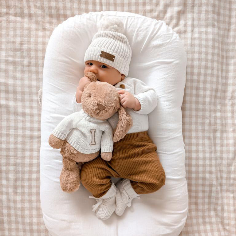 Baby in a white outfit with a brown personalised bunny teddy, lying on a checkered blanket.