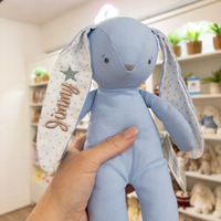 Blue plush Alimrose toy bunny toy with embroidered name 'Jimmy' held by a hand in a store setting.