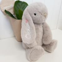 Personalised Jamie Kay Snuggle Bunnies grey beige plush bunny rabbit personalized with name embroidered on it, sitting next to a plant.