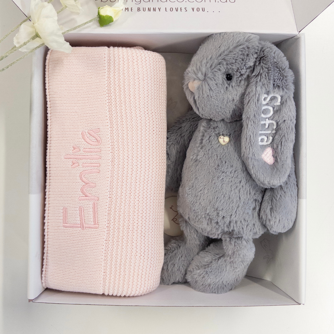 Pink blanket and gray stuffed bunny in a box with 'Emilia' and 'Sofia' embroidered.