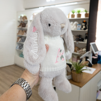 Gray plush bunny toy with embroidered name and design held by a person near rainbow bunny wall