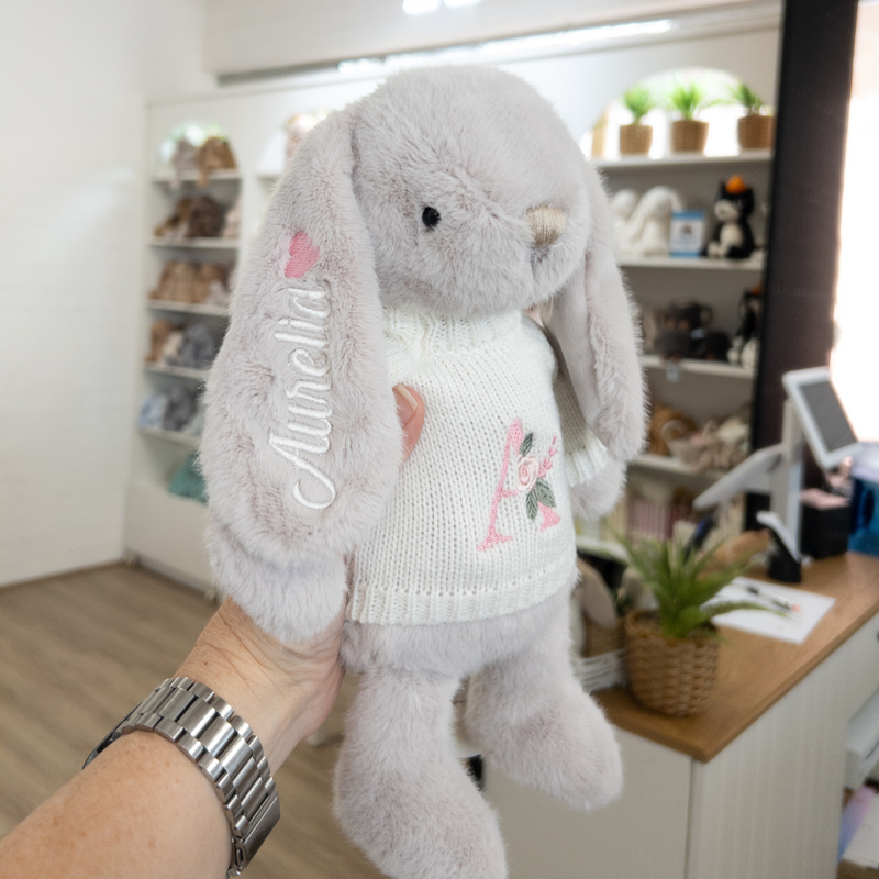 Gray plush bunny toy with embroidered name and design held by a person near rainbow bunny wall