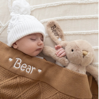 Newborn baby sleeping with a personalised plush bunny rabbit toy and wearing a white knit hat, all on a textured beige personalised baby blanket with diamond knit pattern. Birth announcement photo
