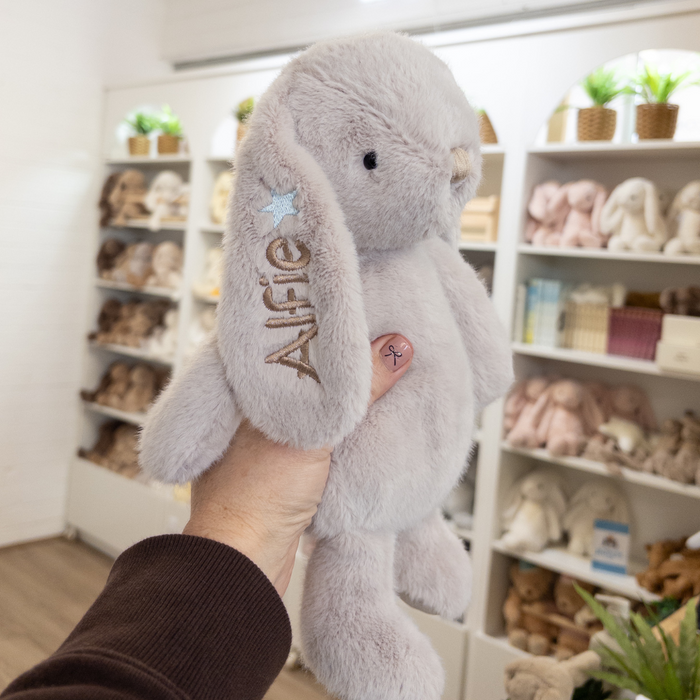 Grey personalised plush bunny rabbit teddy held in a hand with 'Alfie' embroidered on it, in a store setting. Bunny is Jamie Kay Snuggle Bunnies in beige colour for boy or girl gift