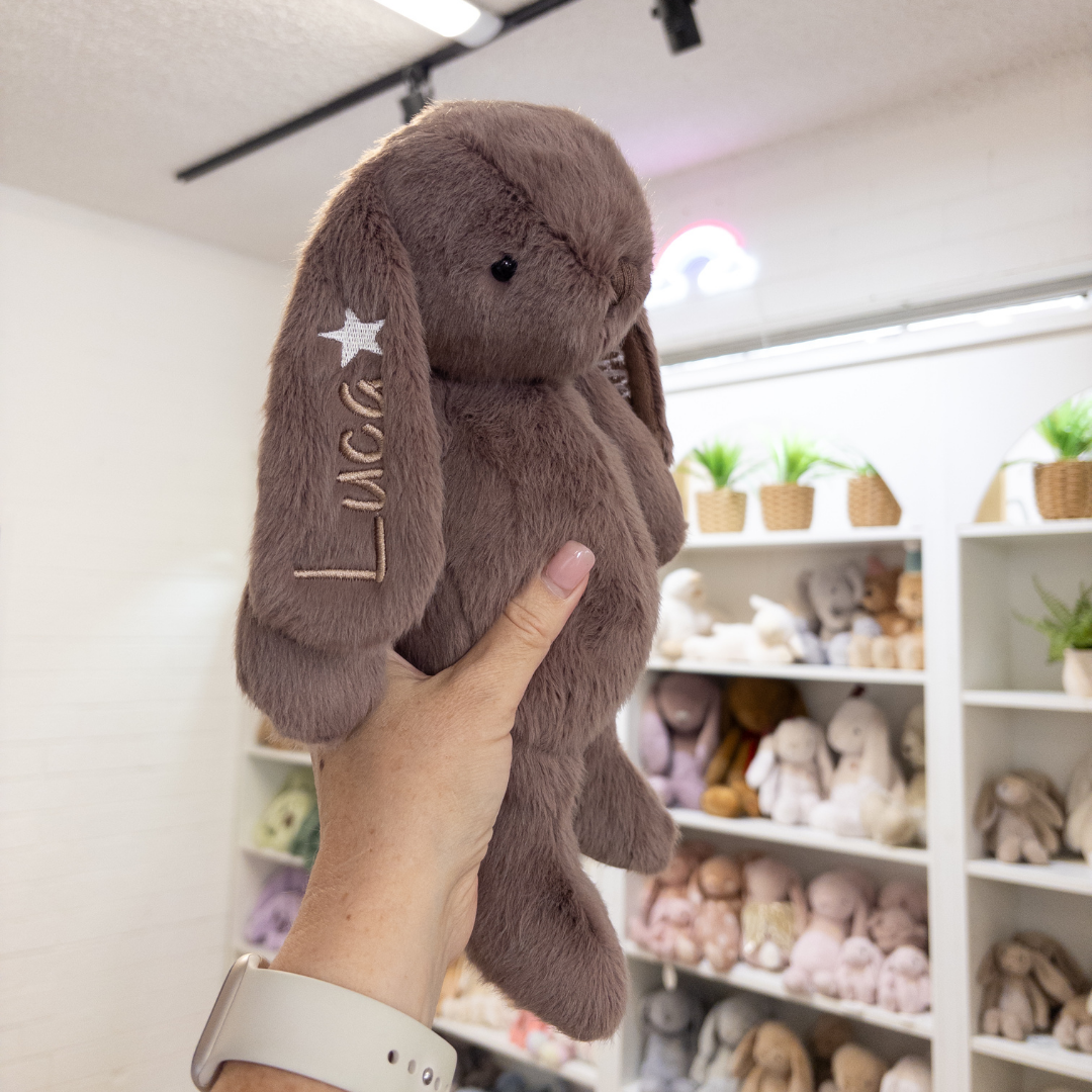 Person holding a brown plush bunny with 'Luca' embroidered on it, in a store setting.