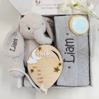 A personalized baby gift set in a white box, featuring a personalised plush light grey elephant teddy, a grey diamond knit blanket, a wooden balloon-shaped birth details card, and a round blue It's a boy cookie on a white background.