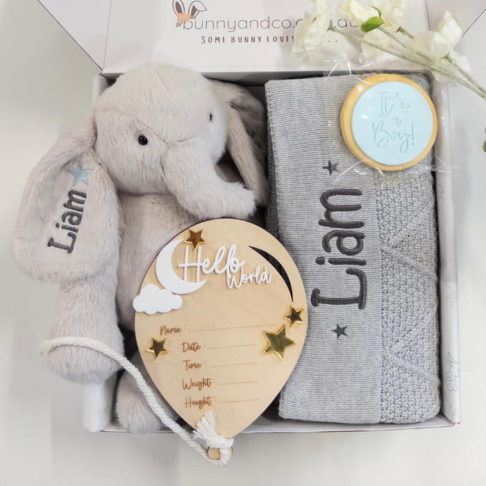 A personalized baby gift set in a white box, featuring a personalised plush light grey elephant teddy, a grey diamond knit blanket, a wooden balloon-shaped birth details card, and a round blue It's a boy cookie on a white background.