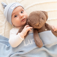 Infant Baby holding a dark brown teddy bunny bear wearing a light blue hat and blanket with 'LUCA' name on it. Jamie Kay Snuggle Bunnies personalised baby gift box australia