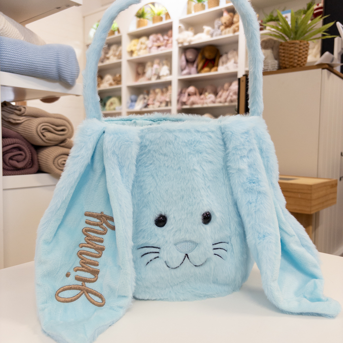 Blue plush bunny easter basket bag with 'Jimmy' embroidered on the ear, sitting on a surface with shelves in the background.